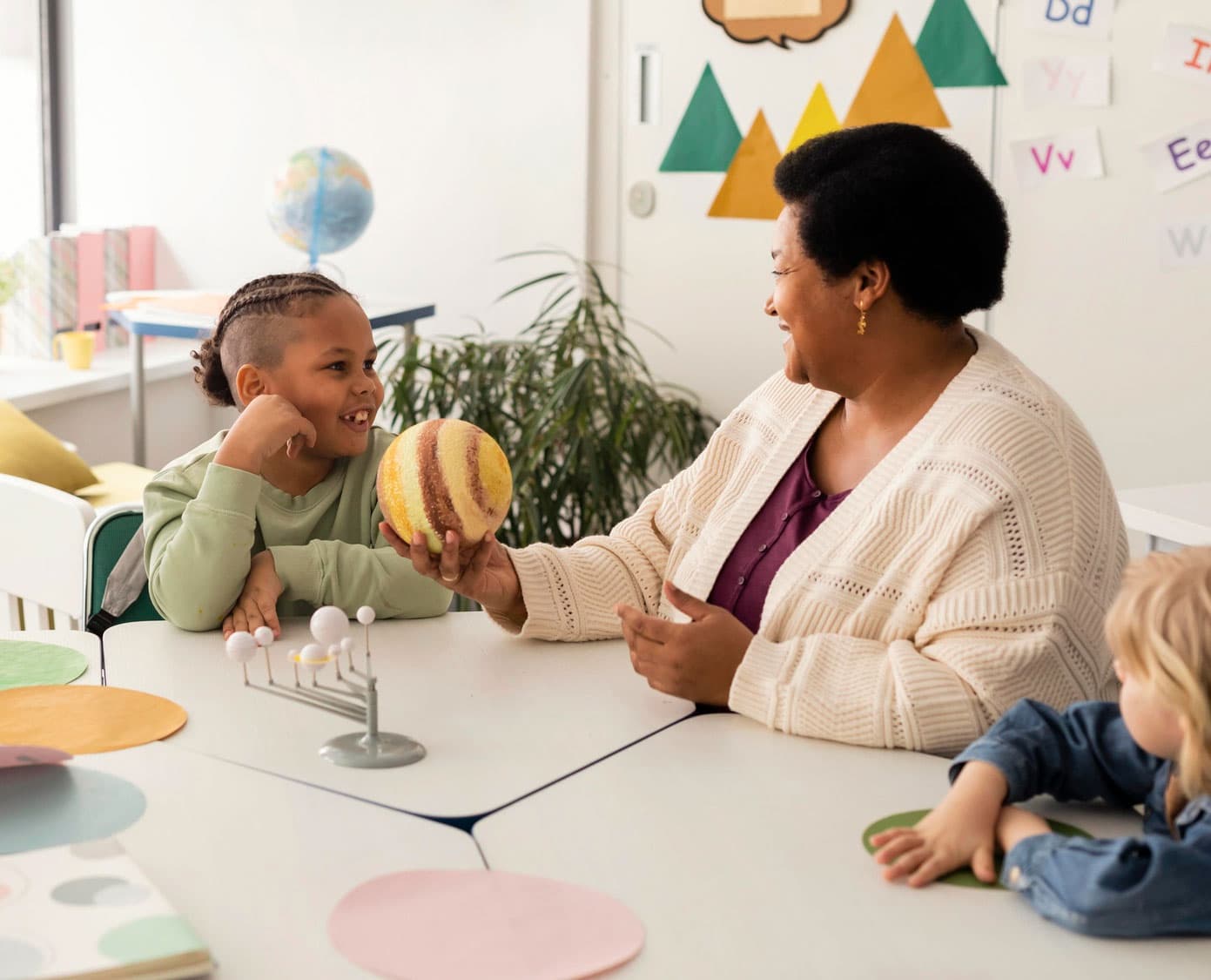 Teacher and a smiling child examining a model planet together beside a small solar-system model