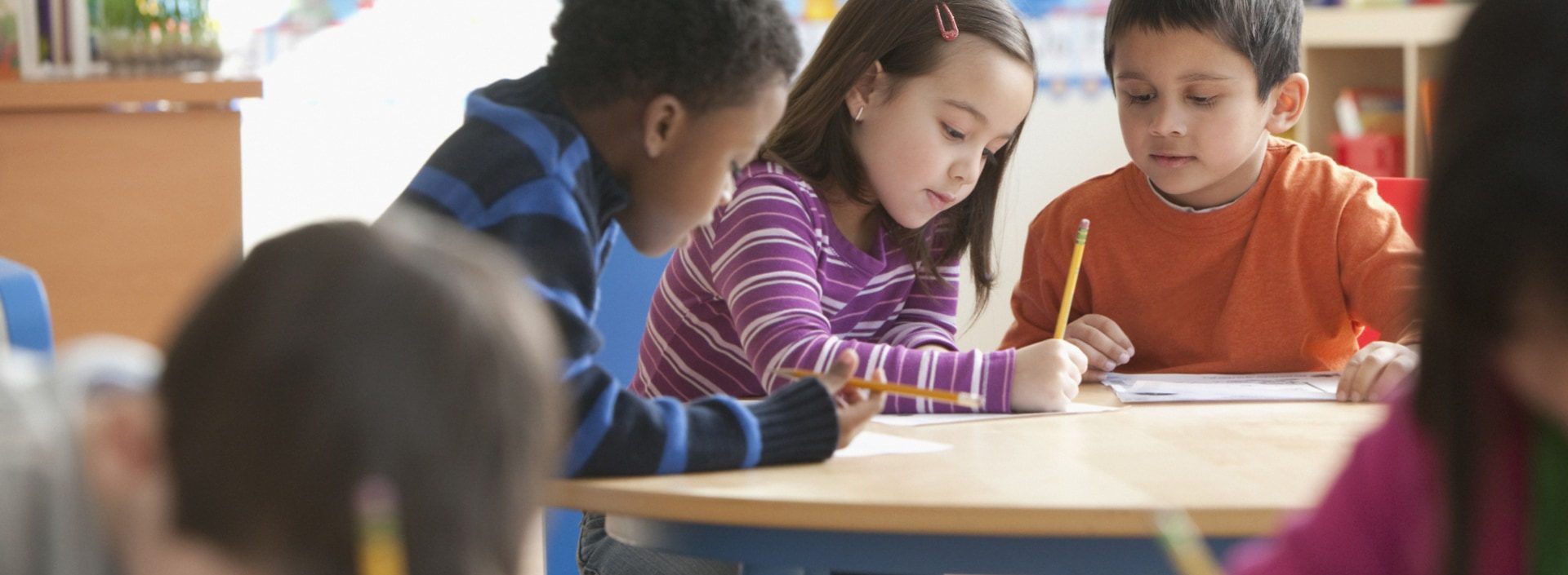 Three children at a classroom table with paper and pencil.